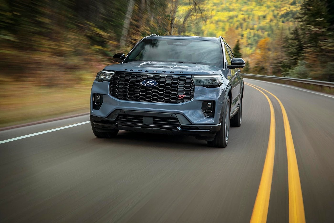 Blue gray 2027 Ford Explorer ST driving a forest highway in fall color, new for Farmington MO buyers