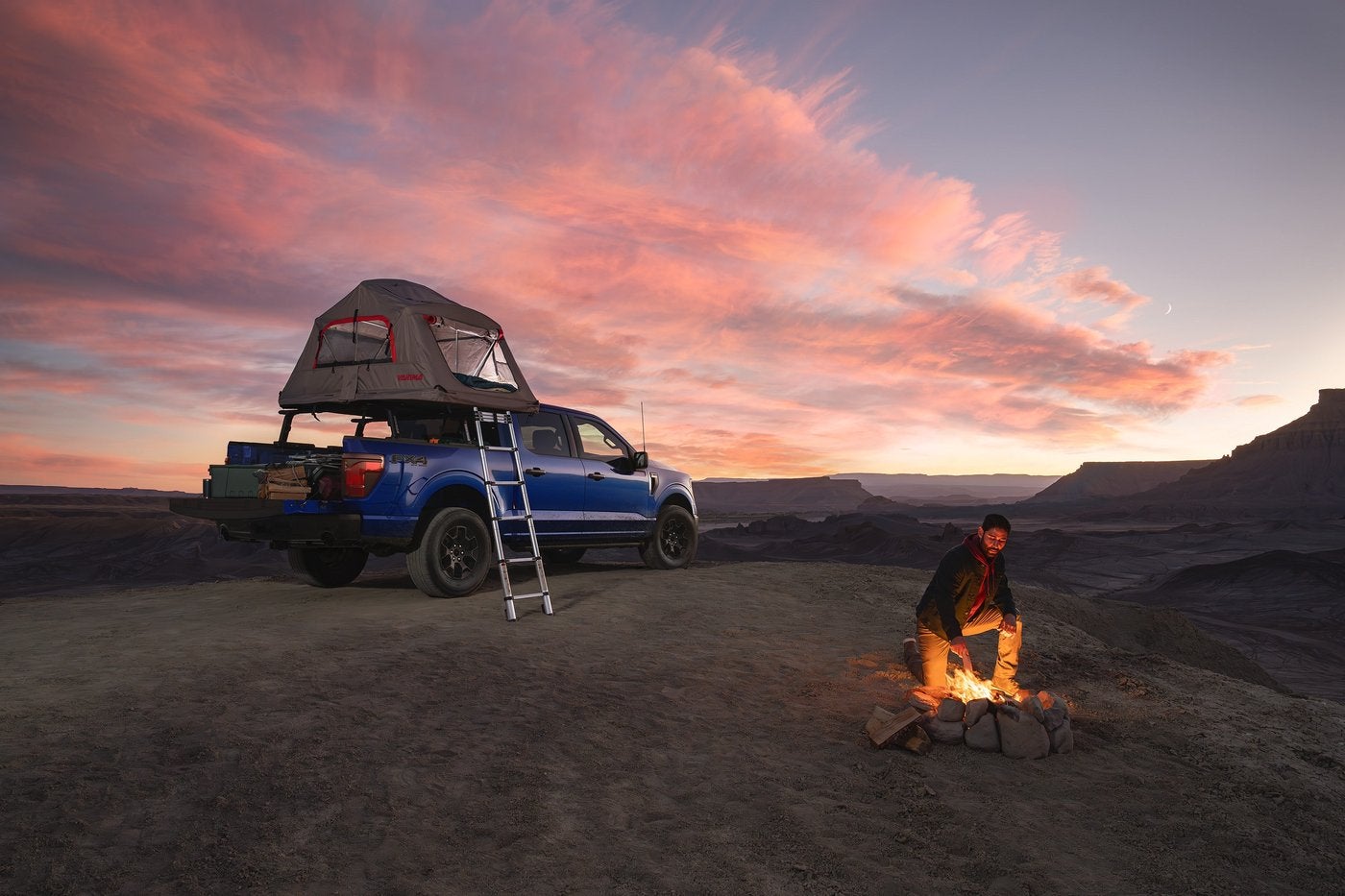 Man starting a campfire beside a blue Ford F-150 with a roof tent at dusk Man starting a campfire beside a blue Ford F-150 with a roof tent at dusk, Farmington MO