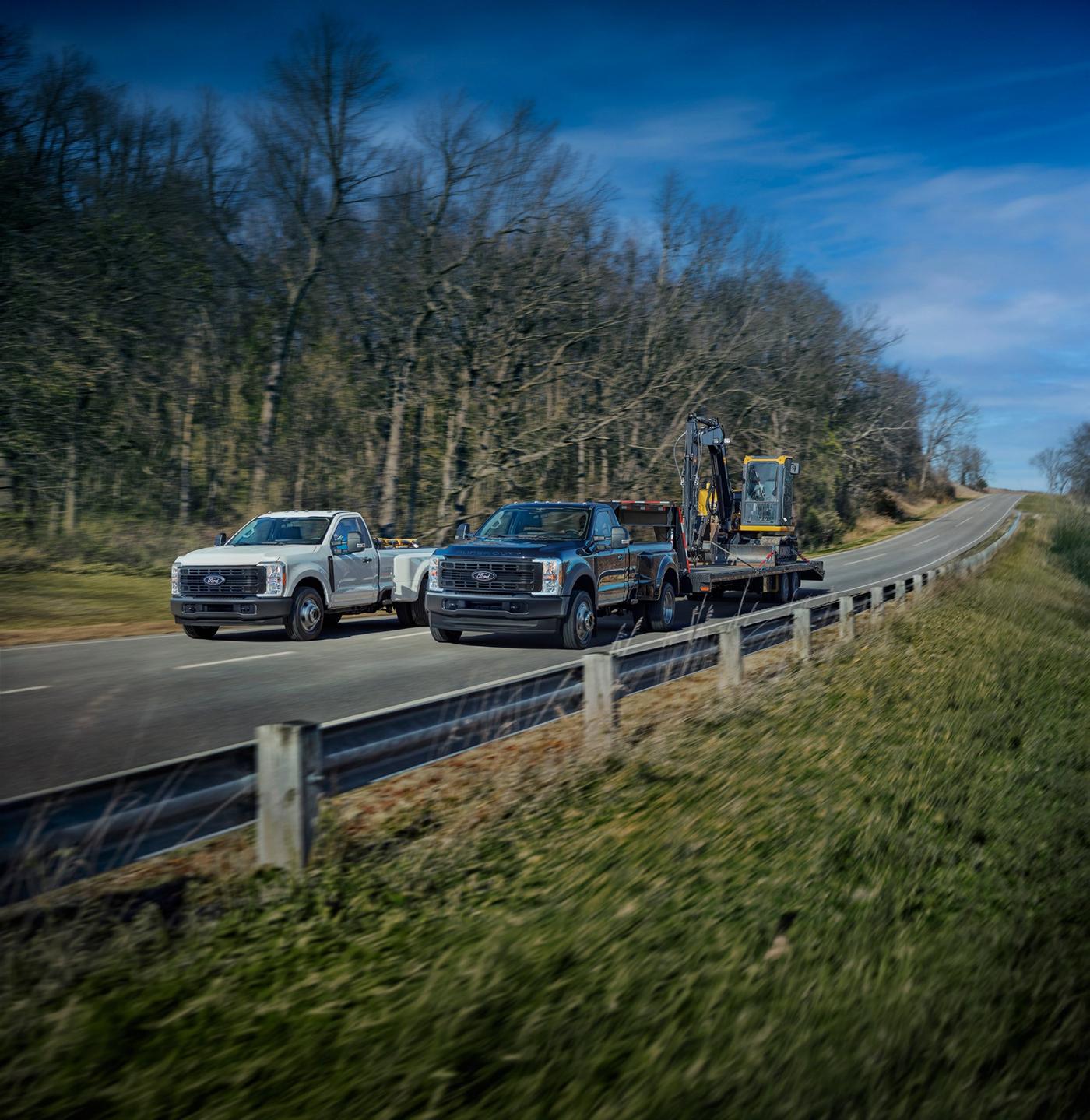 Two Ford Super Duty work trucks on a two lane road, one towing a mini excavator, fleet work around Farmington MO