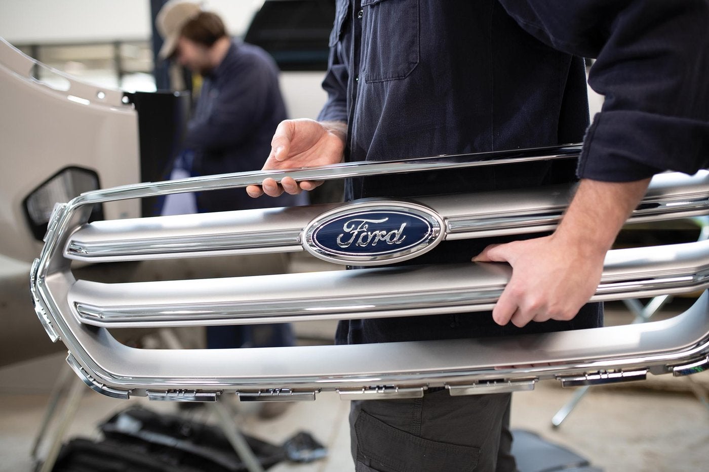 Technician holding a genuine chrome Ford grille with the blue oval in a repair shop, Farmington MO