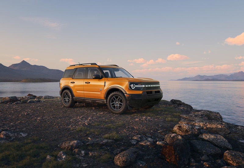Orange Ford Bronco Sport parked on a rocky shore by a lake with mountains behind